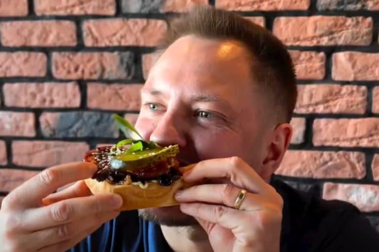 Mężczyzna jedzący kanapkę z mięsem i warzywami w restauracji z ceglastą ścianą w tle.