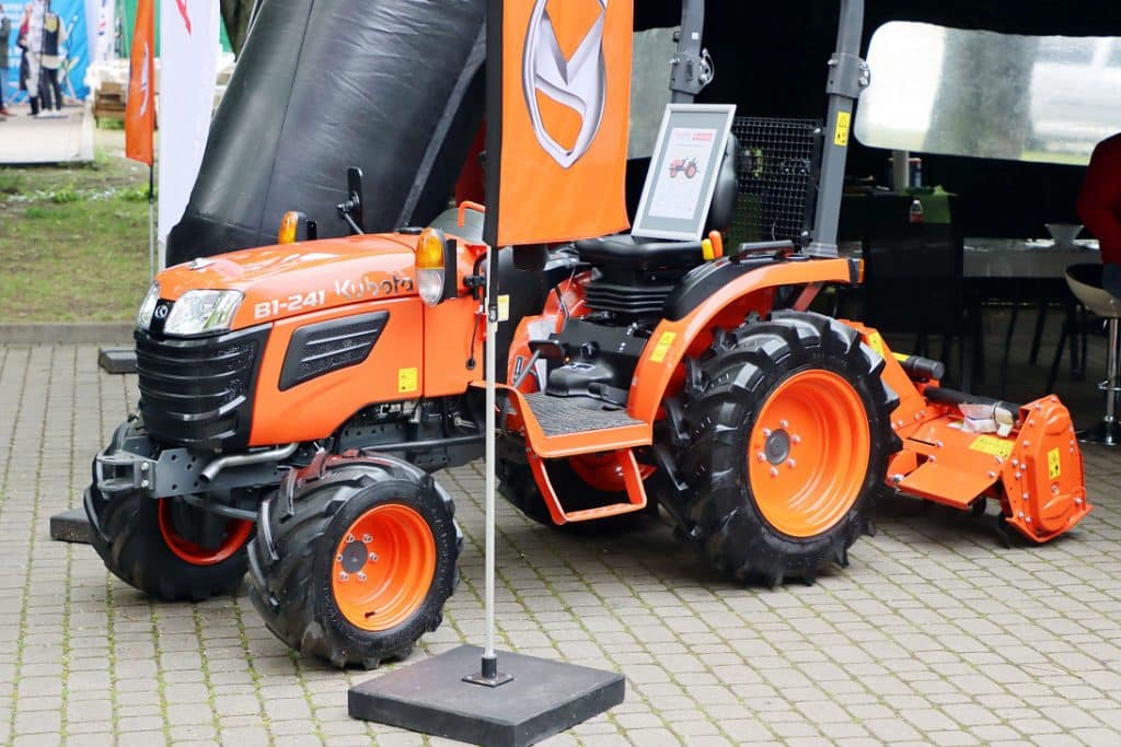 Pomarańczowy traktor na targach ogrodniczych w Olsztynie, ustawiony na brukowanym placu.