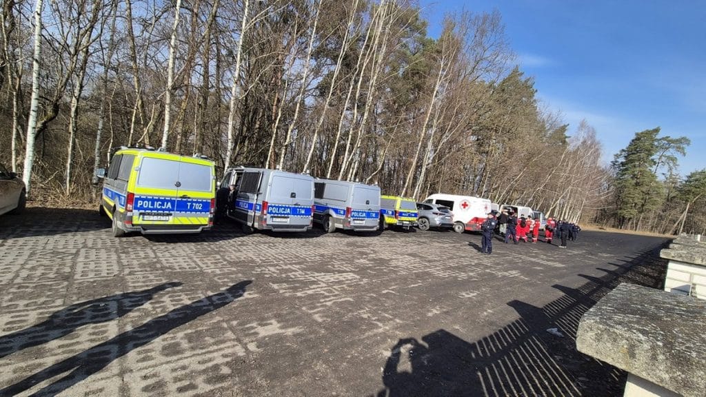 Zesp&oacute;ł służb ratowniczych i policji na miejscu akcji poszukiwawczej w lesie, w pobliżu drogi, w słoneczny dzień.
