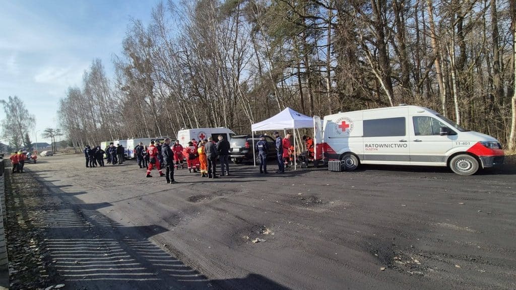 Grupa ratownik&oacute;w i służb poszukiwawczych na terenie lasu w Bartoszycach, z pojazdami i namiotami.