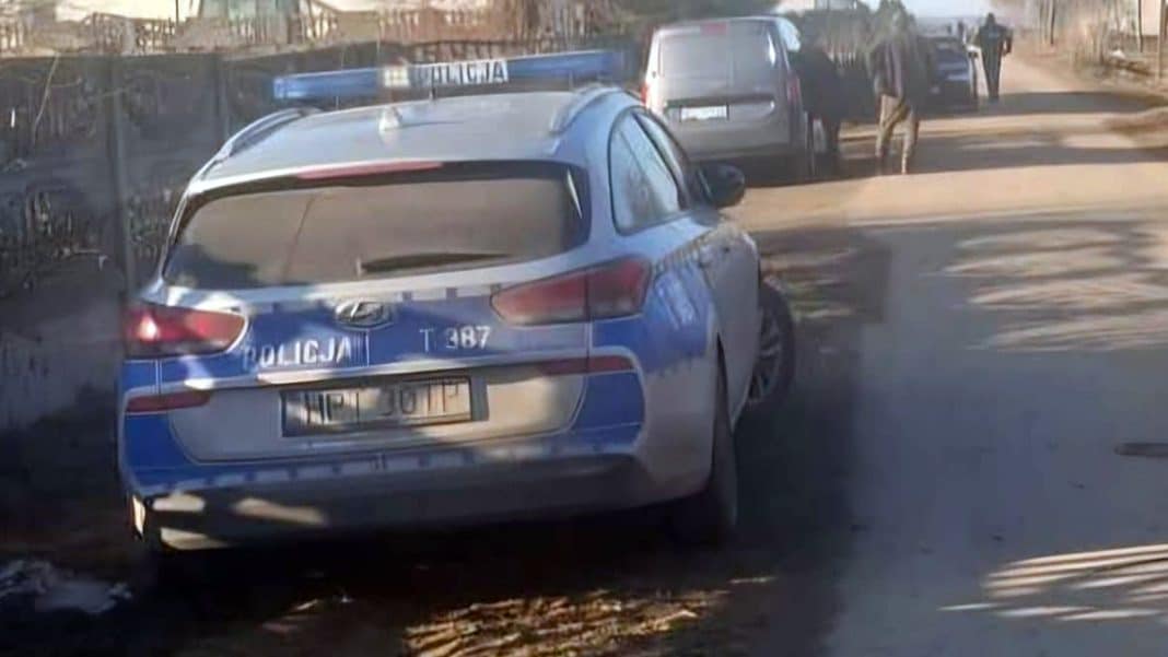 Zdjęcie policyjnego samochodu na wiejskiej drodze, w tle kilka innych pojazd&oacute;w i ludzie.