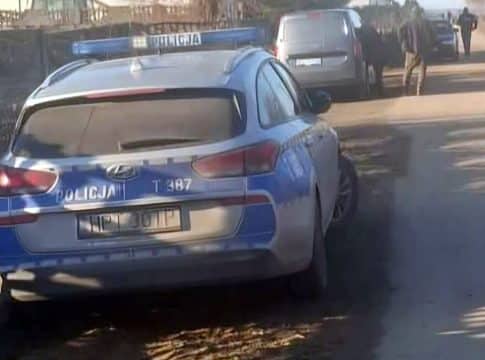 Zdjęcie policyjnego samochodu na wiejskiej drodze, w tle kilka innych pojazd&oacute;w i ludzie.