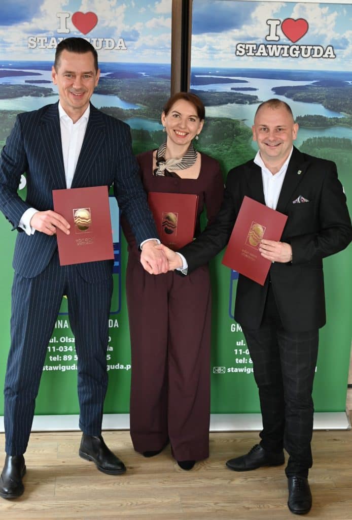 Trzy osoby trzymające dokumenty, uśmiechnięte, na tle mapy i napisu "I &hearts; STAWIGUDA".