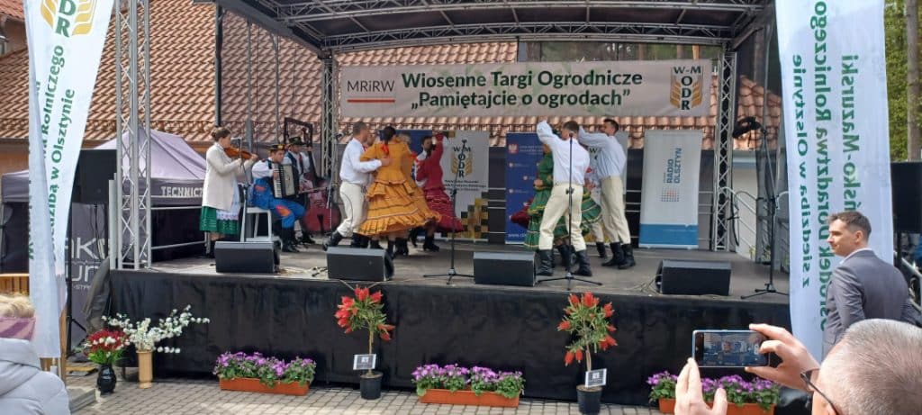 Na scenie występują tancerze w tradycyjnych strojach podczas targ&oacute;w ogrodniczych w Olsztynie.