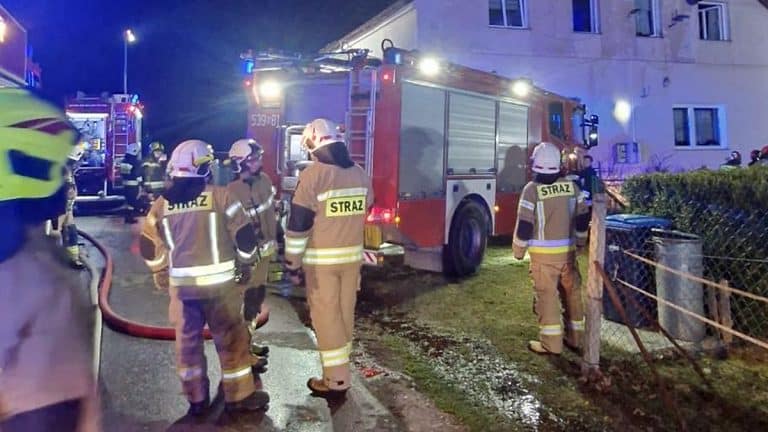 Strażacy gaszą pożar w wielorodzinnym budynku nocą, w tle widoczne są służby ratunkowe i ogień.