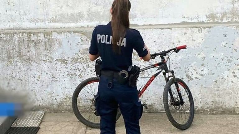 Policjantka z rowerem przed szkołą, widoczna z tyłu, w uniformie, na tle białej ściany.