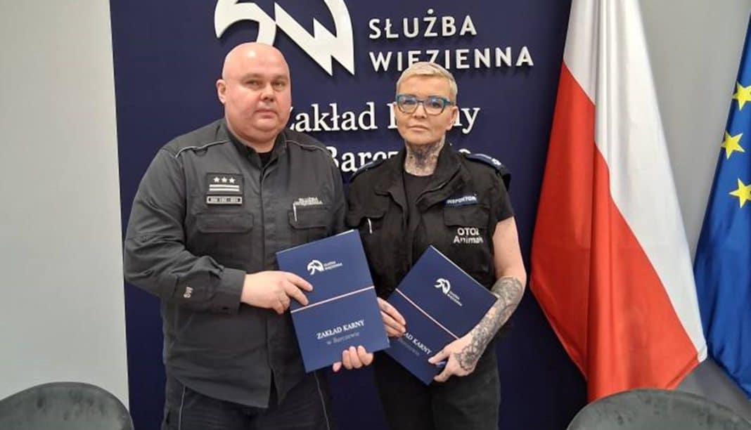 Dw&oacute;ch więźni&oacute;w trzymających dokumenty, stojących przed flagami i tablicą w zakładzie karnym.