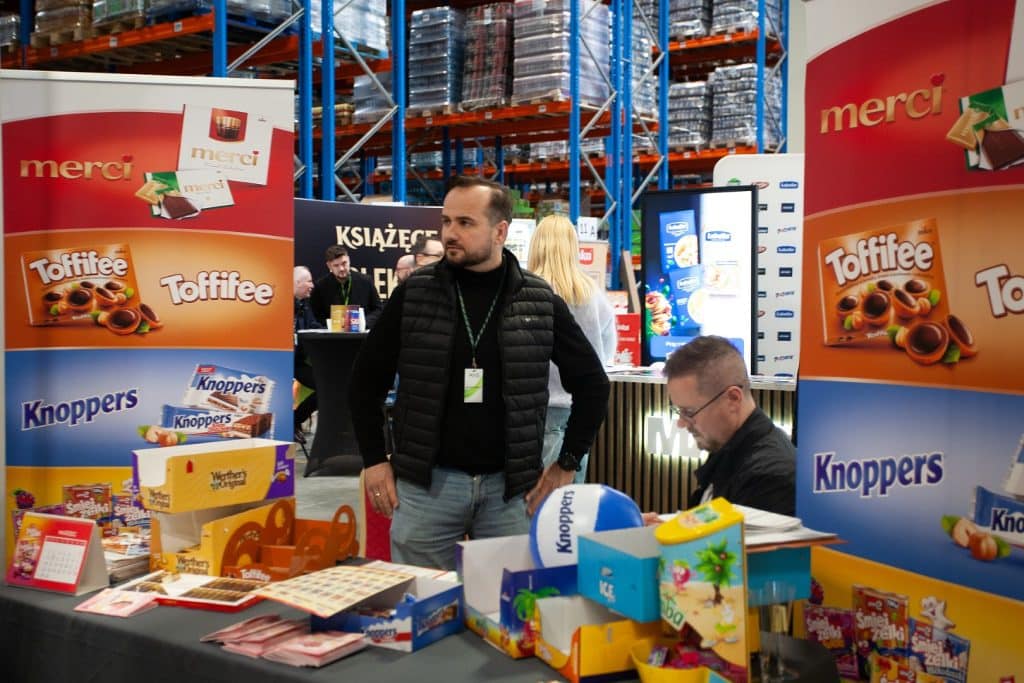 Uczestnicy targ&oacute;w w Gdańsku przy stoisku z produktami marki Merci i Toffifee, w tle regały z towarami.