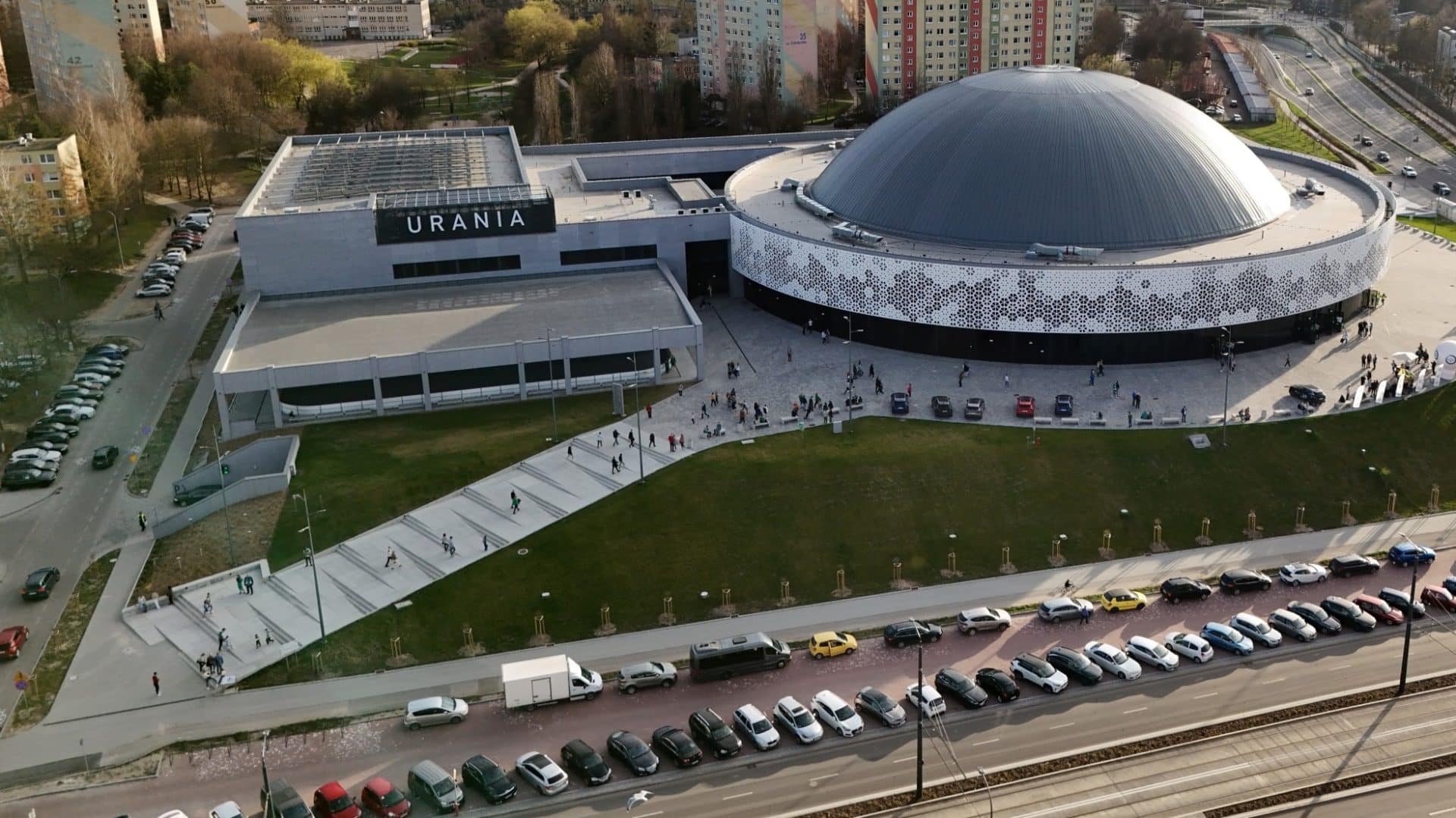 Widok na Halę Urania w Olsztynie z lotu ptaka, otoczona parkingami i zielenią.