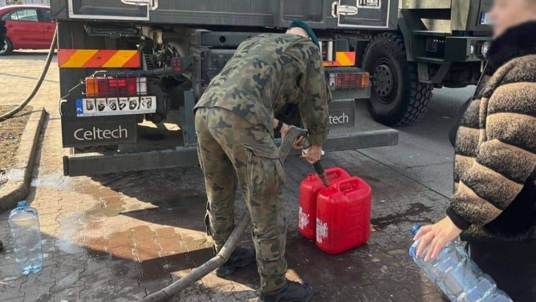 Pracownik z wężem do pompowania wody przy cysternie, obok ludzie z butelkami na wodę.