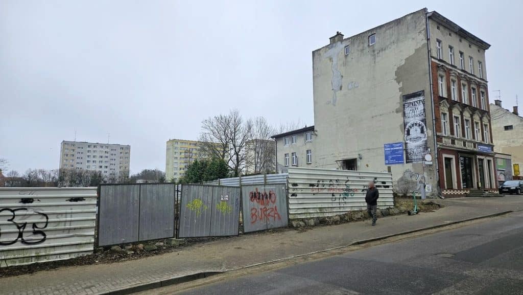 Teren przy szpitalu w Olsztynie, zdemolowany i ogrodzony metalową barierą, w tle stare kamienice i nowoczesne budynki.