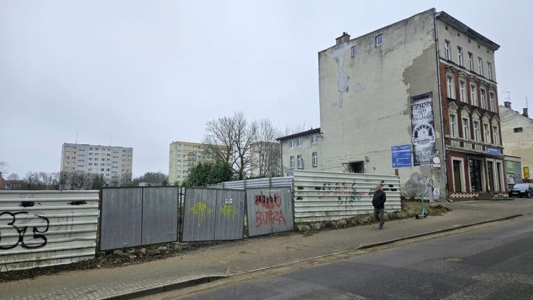 Teren przy szpitalu w Olsztynie, zdemolowany i ogrodzony metalową barierą, w tle stare kamienice i nowoczesne budynki.
