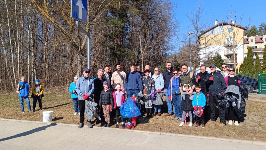 Grupa mieszkańc&oacute;w i uczestnik&oacute;w akcji sprzątania Lasku Pieczewskiego w Olsztynie, zebranych na słonecznym placu.