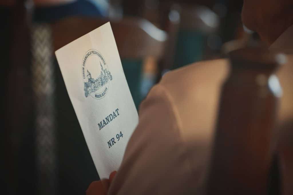 Osoba trzyma dokument z logo i napisem 'Mandat NR 94' podczas wydarzenia w Olsztynie.