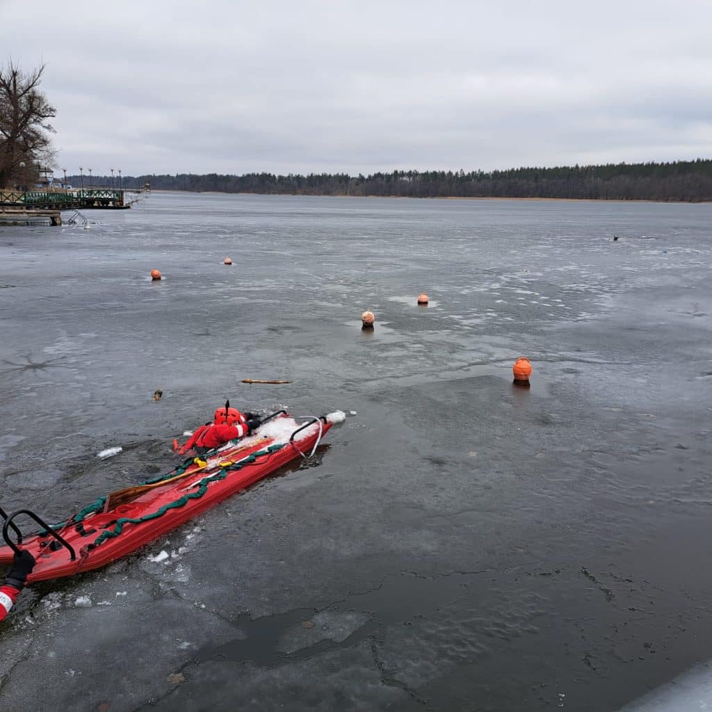 Akcja ratunkowa na zamarzniętym jeziorze, ratownicy w łodzi ratunkowej, w tle zamarznięta powierzchnia.