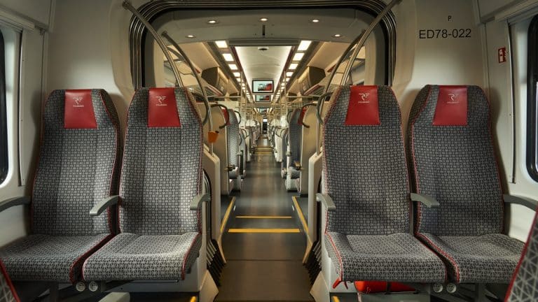 Wnętrze pociągu POLREGIO z pustymi, wygodnymi siedzeniami i ekranem nad przejściem.