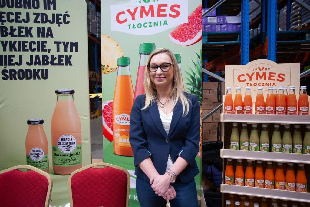 Kobieta w biurze targowym, stojąca obok stoisk z napojami i plakat&oacute;w promocyjnych.