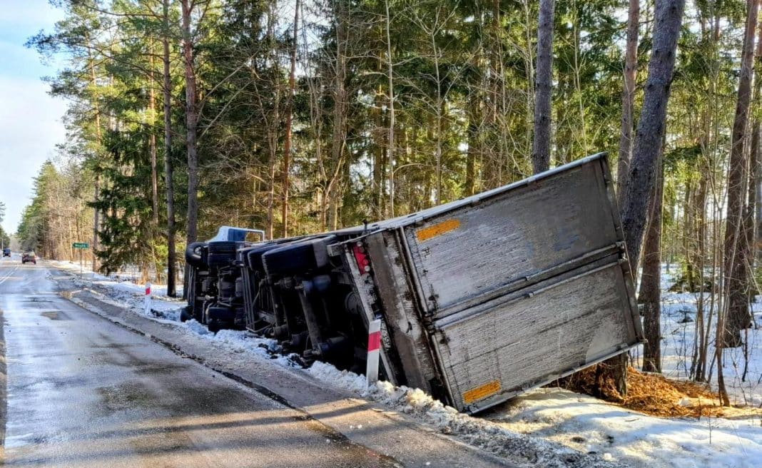 Rozbity samoch&oacute;d ciężarowy leży na boku przy drodze w lesie, wok&oacute;ł śnieg i drzewa.