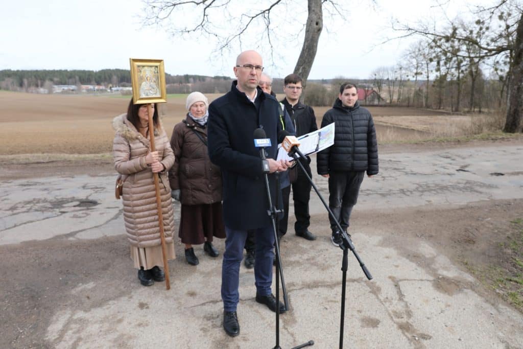Grupa ludzi na zewnątrz, jedna osoba przemawia do mikrofonu, w tle pola i drzewa.