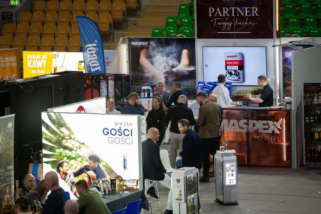 Tłum uczestnik&oacute;w na targach w hali widowiskowo-sportowej, stoiska z produktami i rozmowy biznesowe.