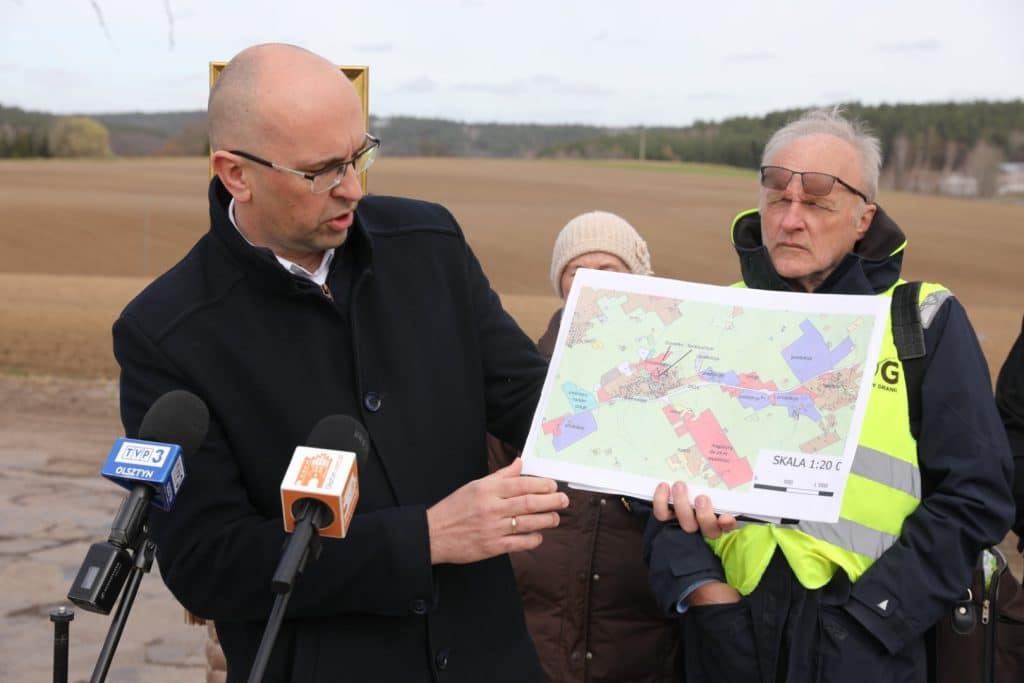 Mężczyzna pokazuje mapę planu zagospodarowania przestrzennego na tle otwartego pola, w tle chmury i krajobraz wiejski.