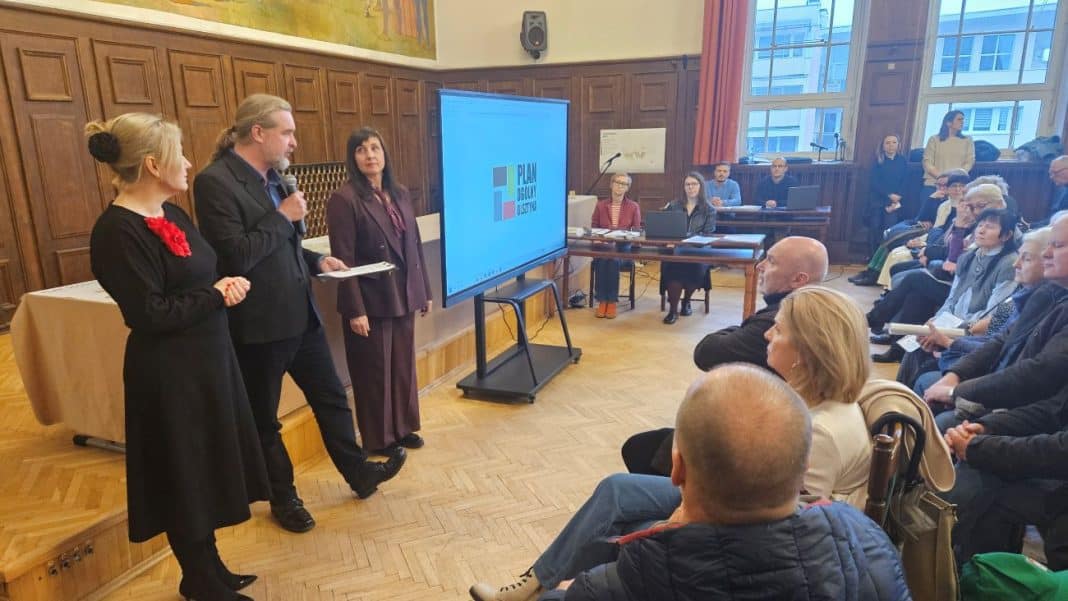 Spotkanie w sali z drewnianymi panelami, ludzie słuchają prezentacji na dużym ekranie.