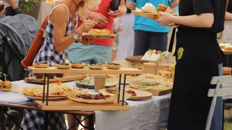 Ludzie na targu degustują potrawy i produkty spożywcze, na stole r&oacute;żnorodne dania i wypieki.