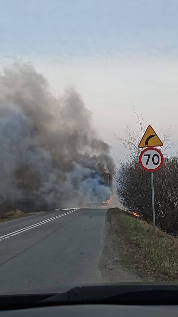 Pożar ciężar&oacute;wki na drodze z widocznym dymem i płomieniami, znak ograniczenia prędkości 70 km/h.