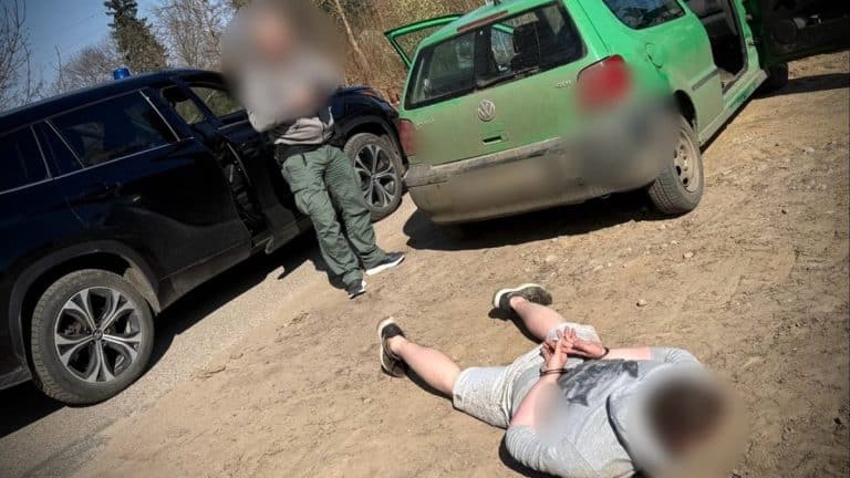 Mężczyzna leży na ziemi z zakładanymi kajdankami, obok dwa samochody, policjant stoi obok.