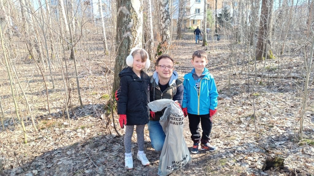 Grupa mieszkańc&oacute;w i dzieci podczas sprzątania lasu w słoneczny dzień, zbierają odpadki.