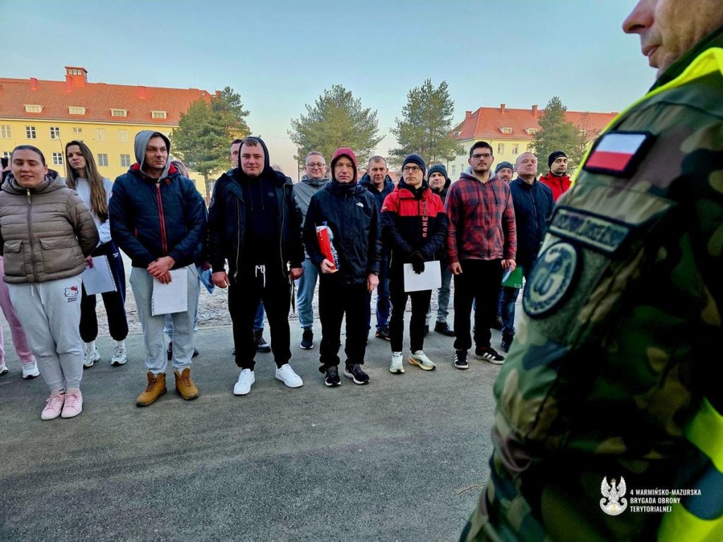 Grupa młodych ludzi stoi na zewnątrz, czekając na rozpoczęcie szkolenia wojskowego, z widokiem na budynki i drzewa.