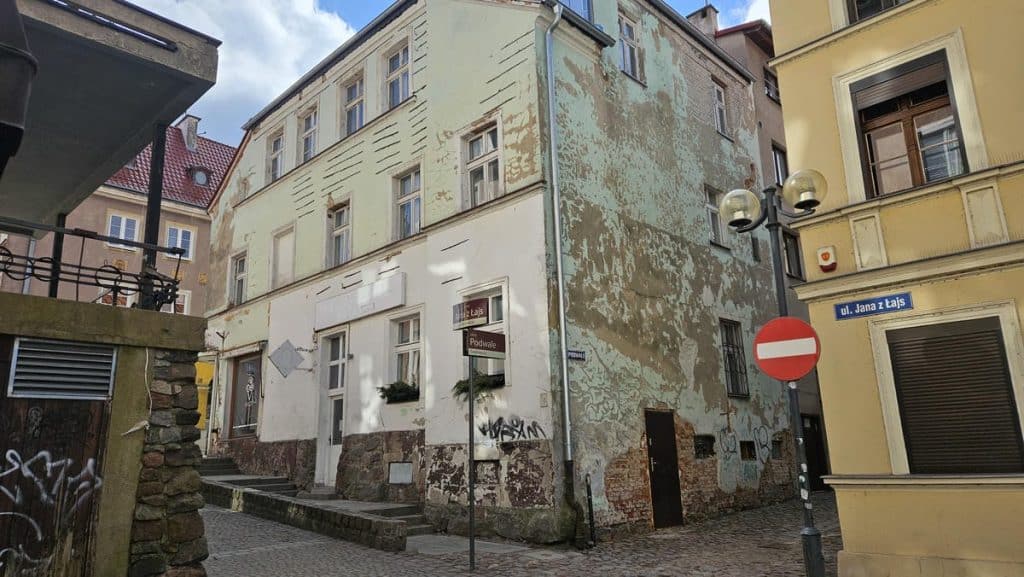 Stara, odrapana kamienica na wąskiej uliczce w centrum Olsztyna, z graffiti na ścianie i znakiem drogowym.