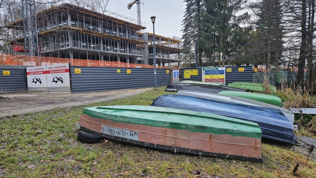 Przed budową nowego obiektu nad jeziorem Ukiel, leżą odwr&oacute;cone kajaki na trawie, w tle plac budowy i ogrodzenie.