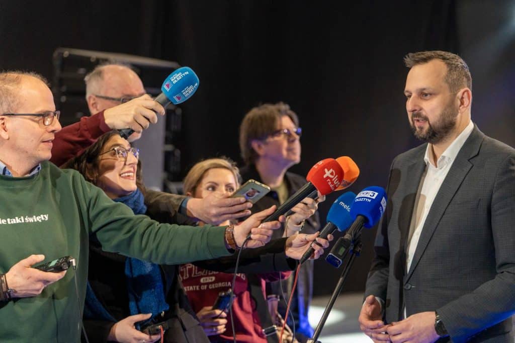 Prezydent Olsztyna podczas konferencji, otoczony dziennikarzami z mikrofonami.