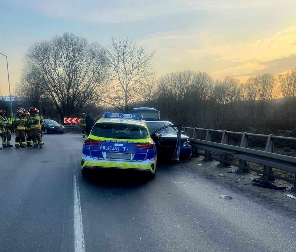 Zdjęcie z wypadku na moście w Nowym Mieście Lubawskim, z radiowozem i rozbitym samochodem, na tle zachodzącego słońca.