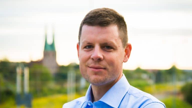 Mężczyzna w niebieskiej koszuli na tle zielonego krajobrazu i wieży kościelnej w oddali.