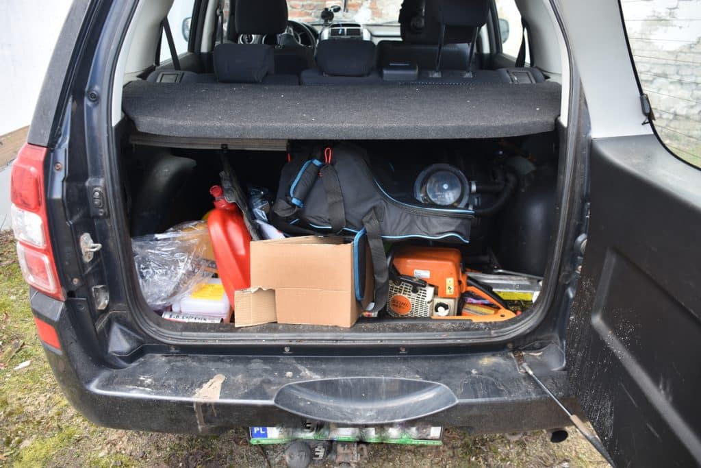 Otwarty bagażnik samochodu z narzędziami i sprzętem, na tle ziemi i ogrodzenia.