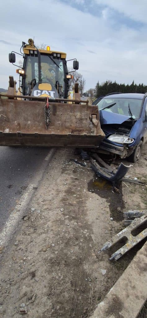 Zdjęcie zderzenia samochodu z koparką na drodze, widoczne uszkodzenia pojazd&oacute;w i służby na miejscu.