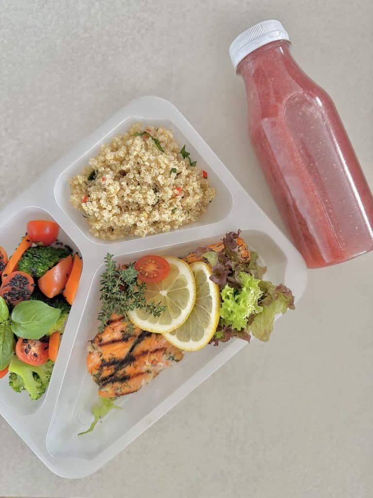 Zdjęcie zdrowego posiłku w pudełku, zawierającego łososia, sałatę, pomidorki, cytrynę, quinoa i smoothie.