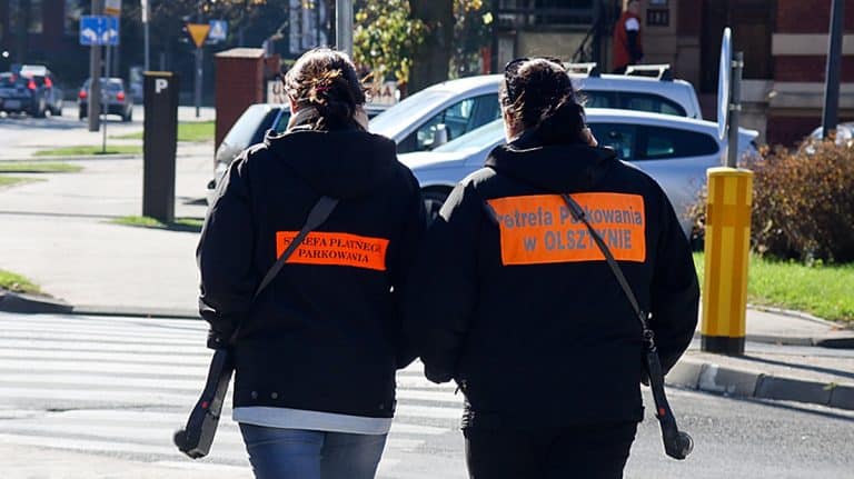 Dwie osoby w czarnych kurtkach z pomarańczowymi napami, patrolujące strefę parkowania w Olsztynie.