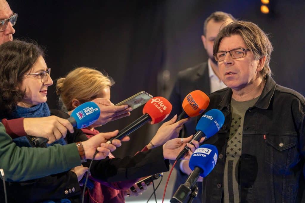 Mężczyzna w okularach i czarnej kurtce udziela wywiadu, otoczony mikrofonami od r&oacute;żnych medi&oacute;w.