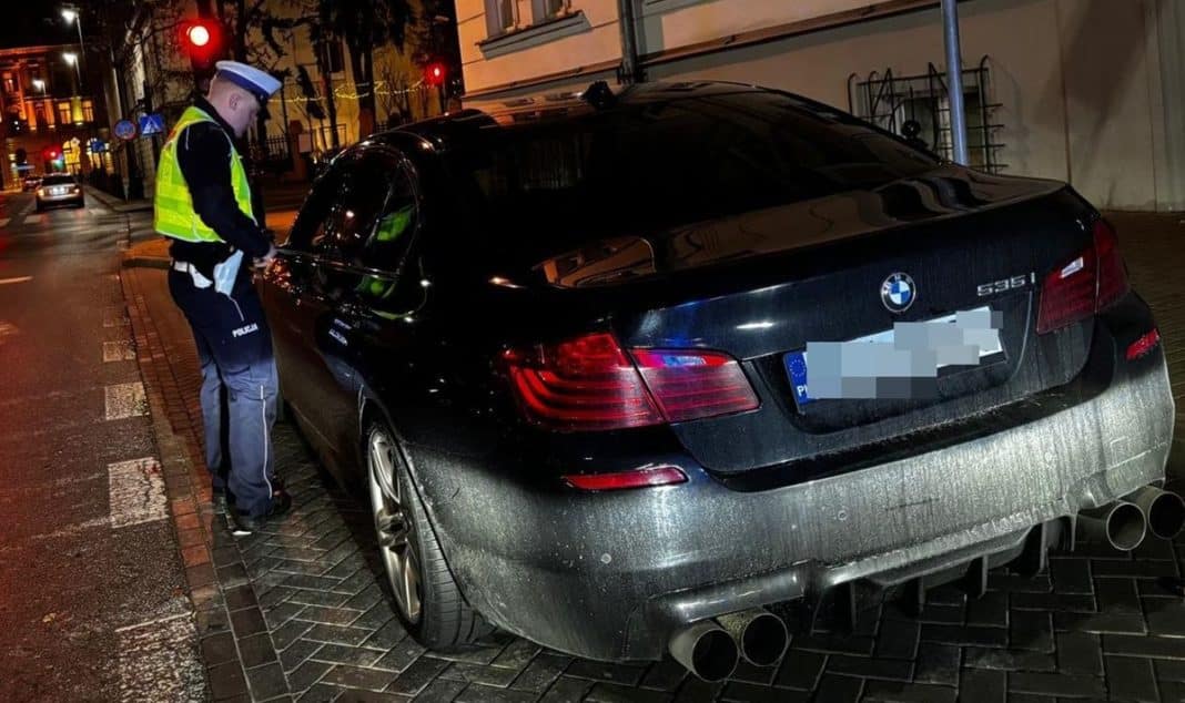 Policjant sprawdza samoch&oacute;d BMW na parkingu po nocnym pościgu, w tle ulice miasta oświetlone latarniami.