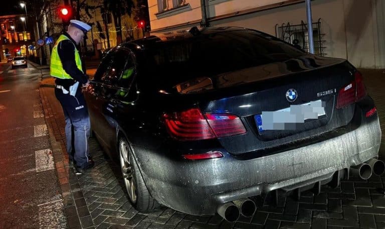 Policjant sprawdza samoch&oacute;d BMW na parkingu po nocnym pościgu, w tle ulice miasta oświetlone latarniami.
