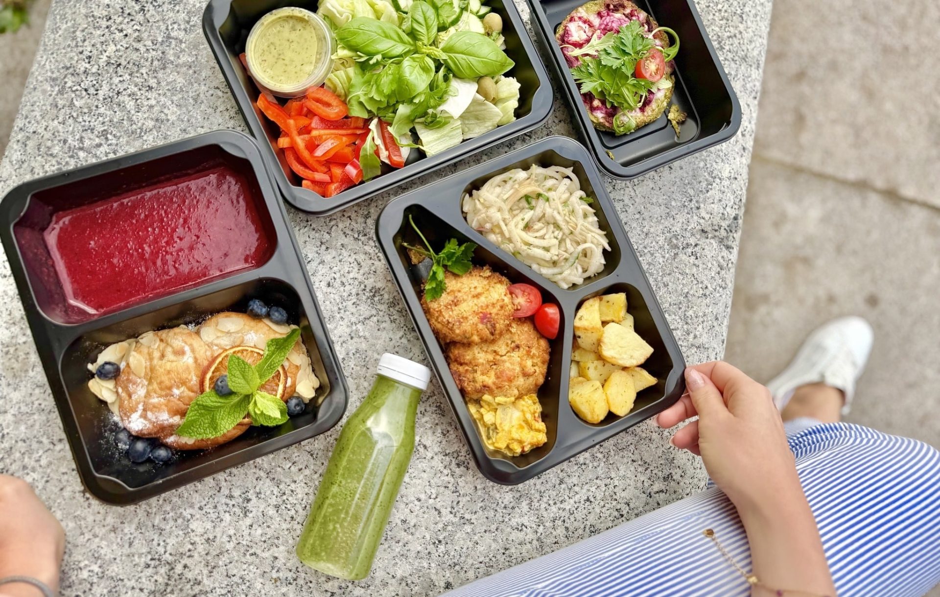 Zestaw dań bezglutenowych w pudełkach na kamiennym stole, obok butelka zielonego napoju.