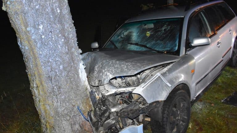 Rozbity samoch&oacute;d po wypadku z uszkodzonym przodem i uderzeniem w drzewo, nocą na drodze.