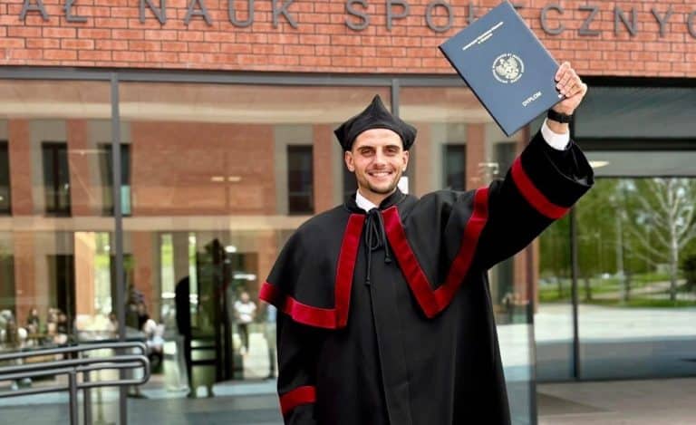 Uczestnik w togach akademickich trzyma dyplom przed budynkiem uczelni.