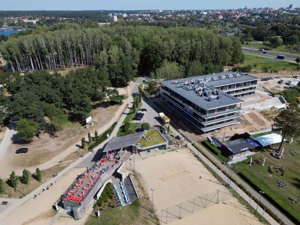 Widok na jezioro Ukiel w Olsztynie, otoczone lasami i nowoczesnymi budynkami, z placem i infrastrukturą rekreacyjną.