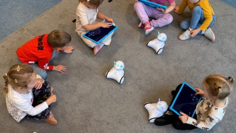 Dzieci siedzą na dywanie, bawiąc się robotami i tabletami w przedszkolu.