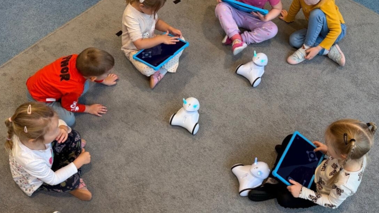 Dzieci siedzą na dywanie, bawiąc się robotami i tabletami w przedszkolu.