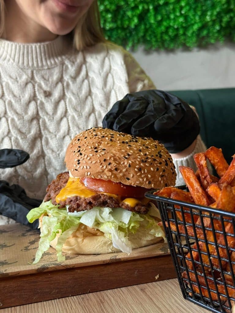 Burger z szarpaną wołowiną, sałatą, pomidorem i serem, podany na drewnianym talerzu obok frytek.